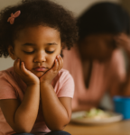 Young girl frowns over an inadequate meal. In the background, her mother holds her had in her hands.