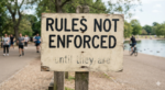 A weathered sign on a beach, with people walking by. In large visible letters, "RULES NOT ENFORCED". Then, underneath in smaller letters barely visible from weather,, "until they are"