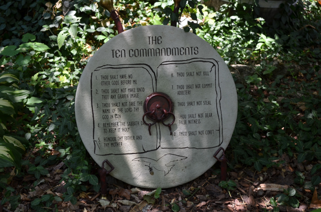 Circular shaped stone with the Ten Commandments chiseled into it, leaning against a green bush.